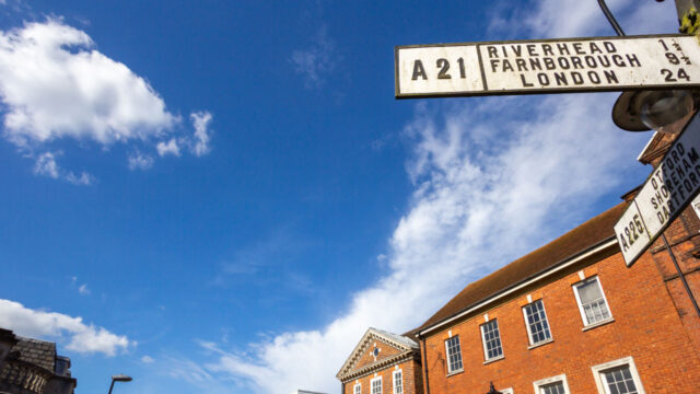 An angled view of a street sign that says "Riverhead Farnborough London" and brick buildings in the background