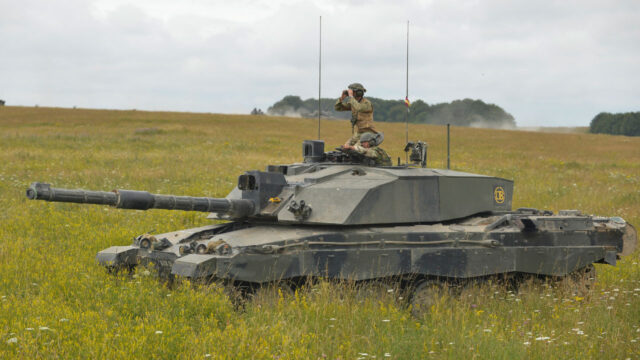 Person in army uniform using binoculars riding on tank in field