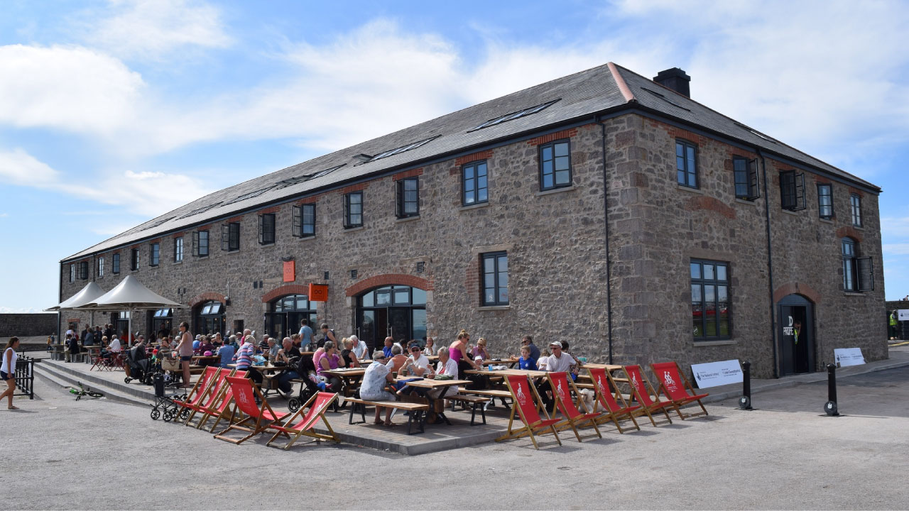 Outdoor dining area with picnic tables, umbrellas, and lounge chairs by stone building