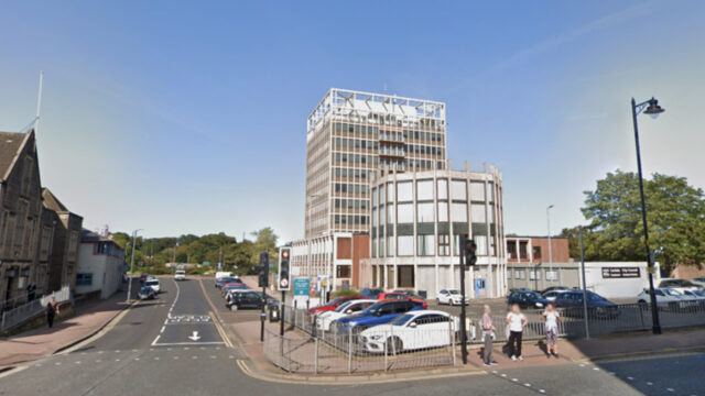 Multi-story building with car park, street parking, and people at crosswalk