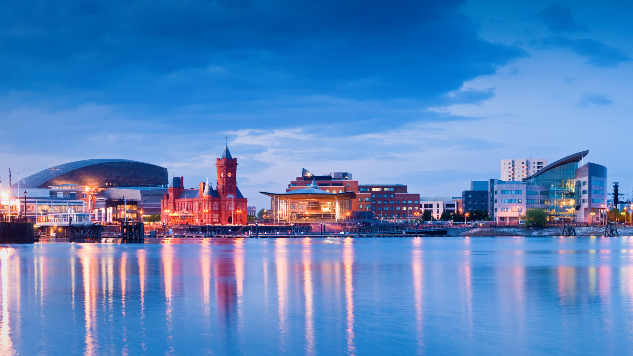 Brightly lit city buildings on water with blue sky in the background and lights reflecting on the water