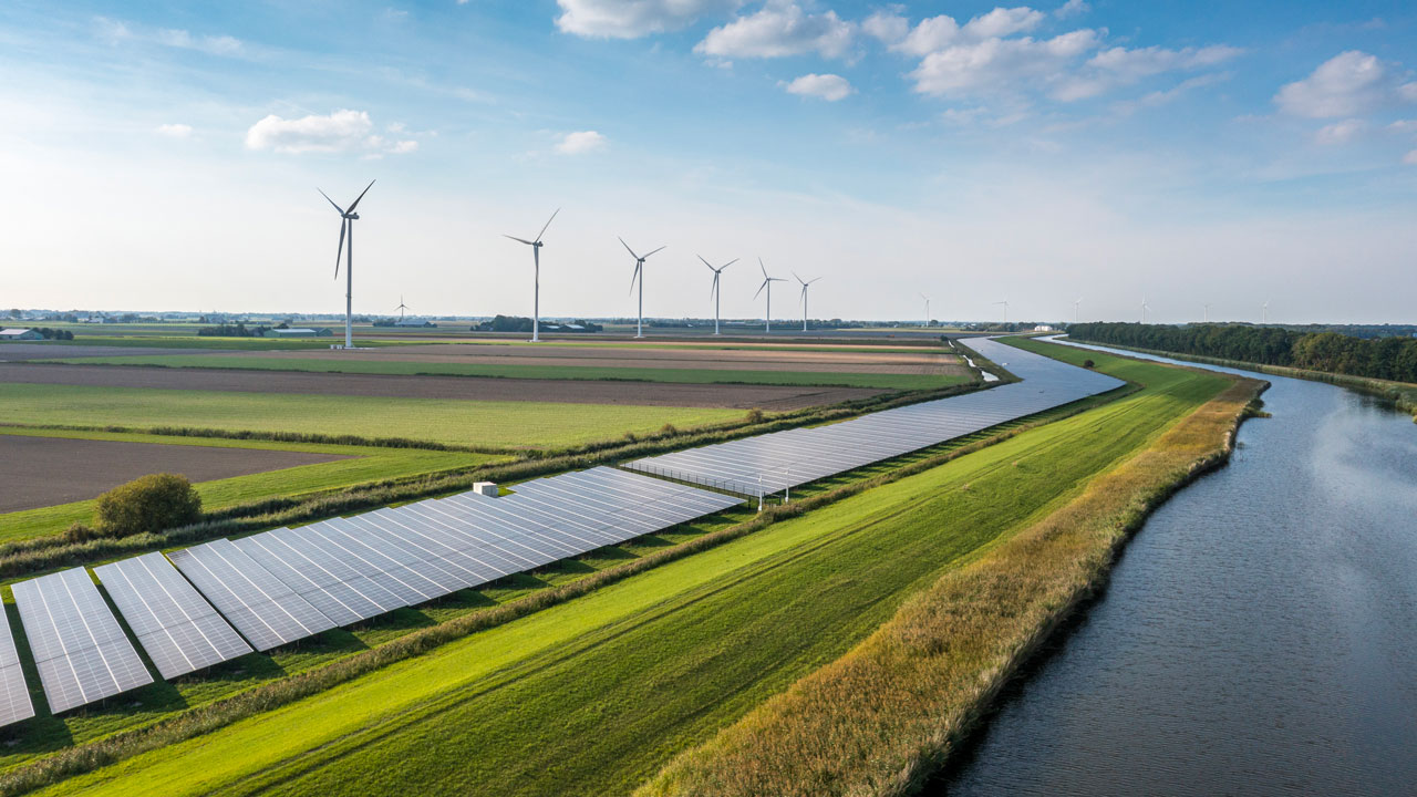 Solar panels lining a winding river with wind turbines in the background