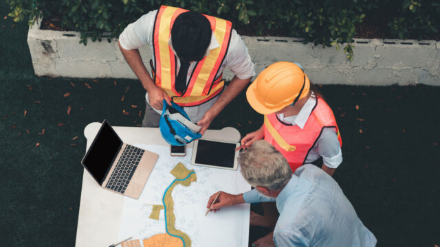 three people gather on a table with plans and a laptop. They are wearing safety clothing