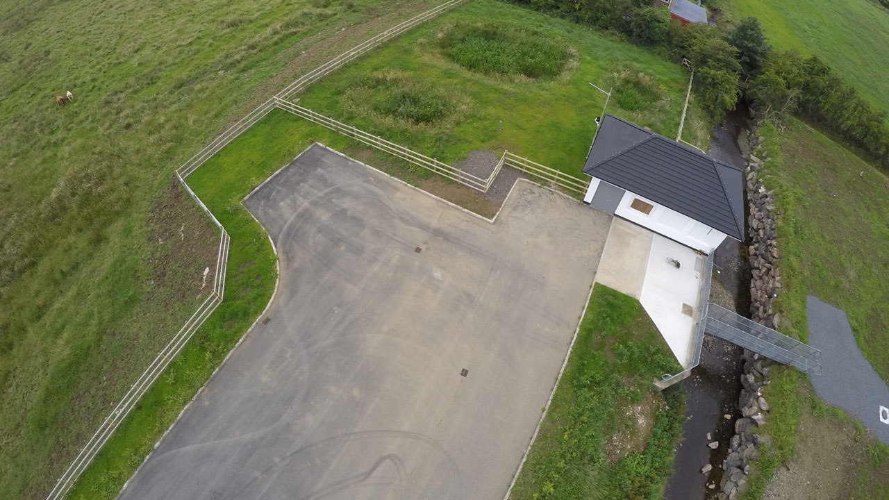 Angled aerial view of a small white wastewater treatment facility, an expanse of pavement in front of it and the houses in the village in the background