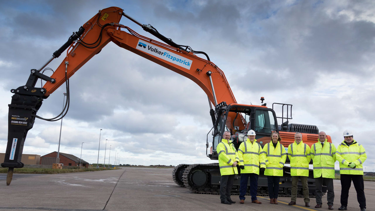 Six personnel in PPE on paved runway in front of heavy machinery