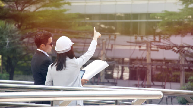 A consultant in a white hard hat standing with a client in a suit, holding documents in one hand and pointing at the building in front of them with another hand