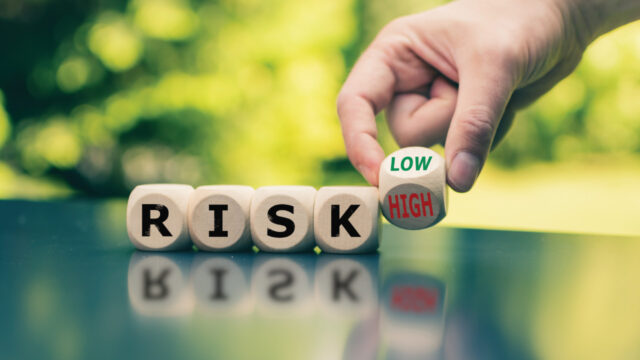 Four wooden dice that spell out the word risk sit on a surface with a hand holding another dye that reads low/high reaching down from above