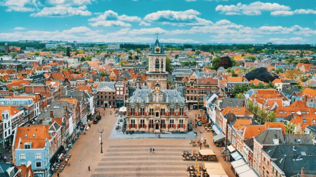 Aerial view of a charming Dutch town square featuring a historic building with a clock tower at its center. Surrounding the square are rows of traditional houses with orange rooftops, interspersed with trees and small market stalls. The sky is bright blue with fluffy white clouds, and the scene is lively with people and bicycles.